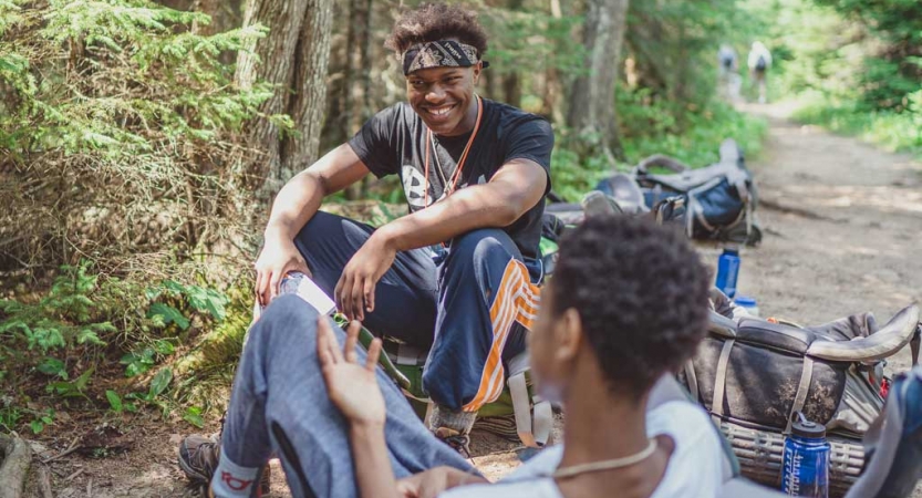 Two people sit on the ground in a wooded area with their backpacks nearby